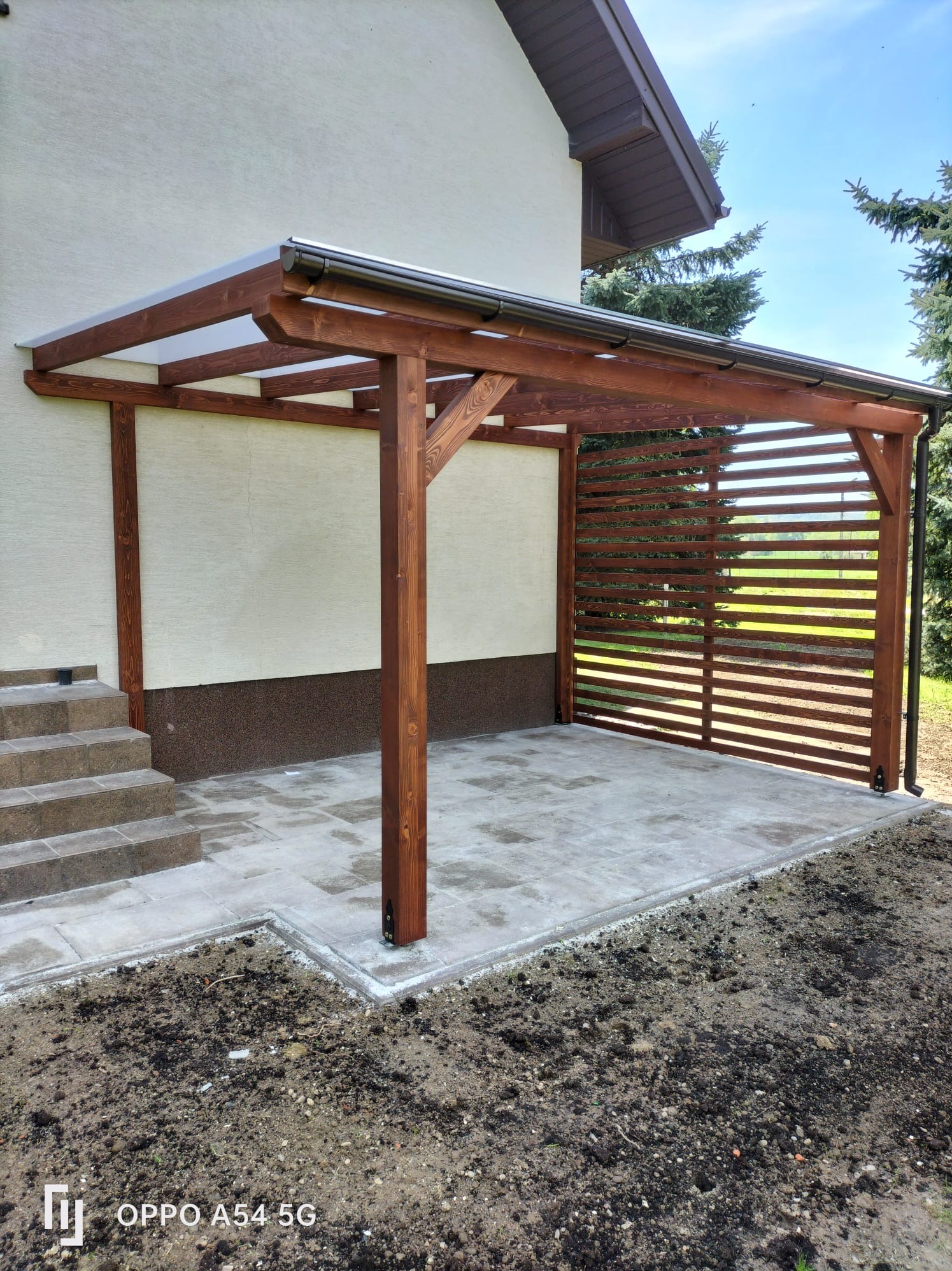 Drewniana pergola z zadaszeniem z poliwęglanu, przylegająca do budynku, z ażurową ścianą i betonową posadzką. Widoczne schody wejściowe i fragment ogrodu.