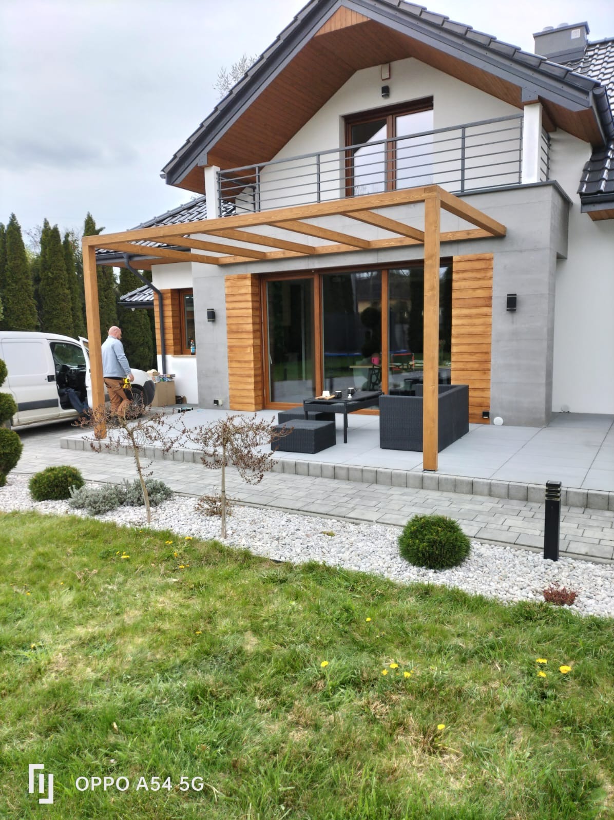 Nowoczesna pergola z jasnego drewna przy domu z tarasem i meblami ogrodowymi. Widoczny pracownik i bus dostawczy. Trawa, krzewy i kamień w ogrodzie.