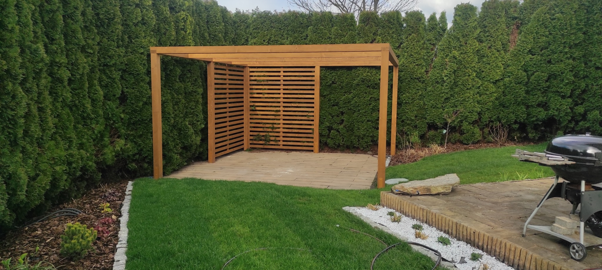 Drewniana pergola z ażurowymi ściankami, otoczona zielenią w ogrodzie. Kamienna podłoga i grill w tle. Ujęcie z niskiej perspektywy.