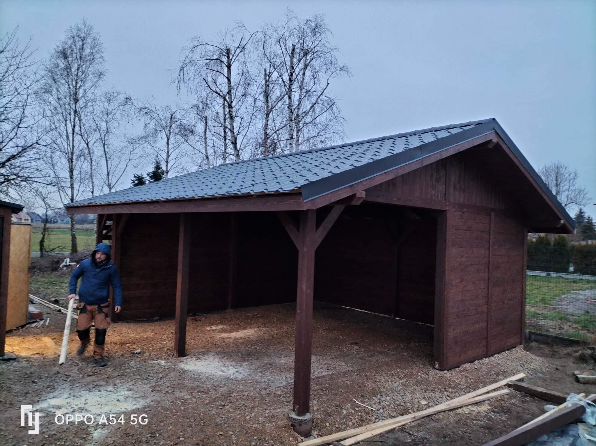 Drewniana wiata z szarym dachem na żwirowym podłożu, widok z zewnątrz. Mężczyzna w roboczym ubraniu niesie drewniany element. Konstrukcja z ciemnego drewna.