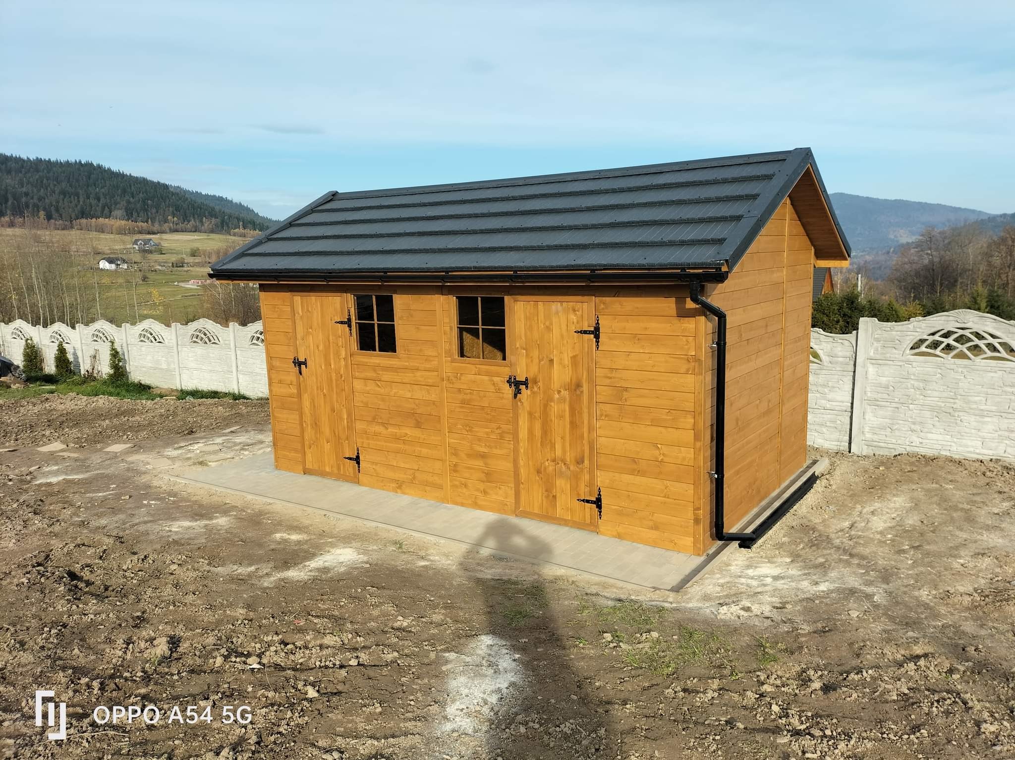 Drewniana altana ogrodowa z czarnym dachem, widoczna w Mszanie Dolnej. Na pierwszym planie świeżo przygotowany teren pod trawnik. W tle góry i błękitne niebo.