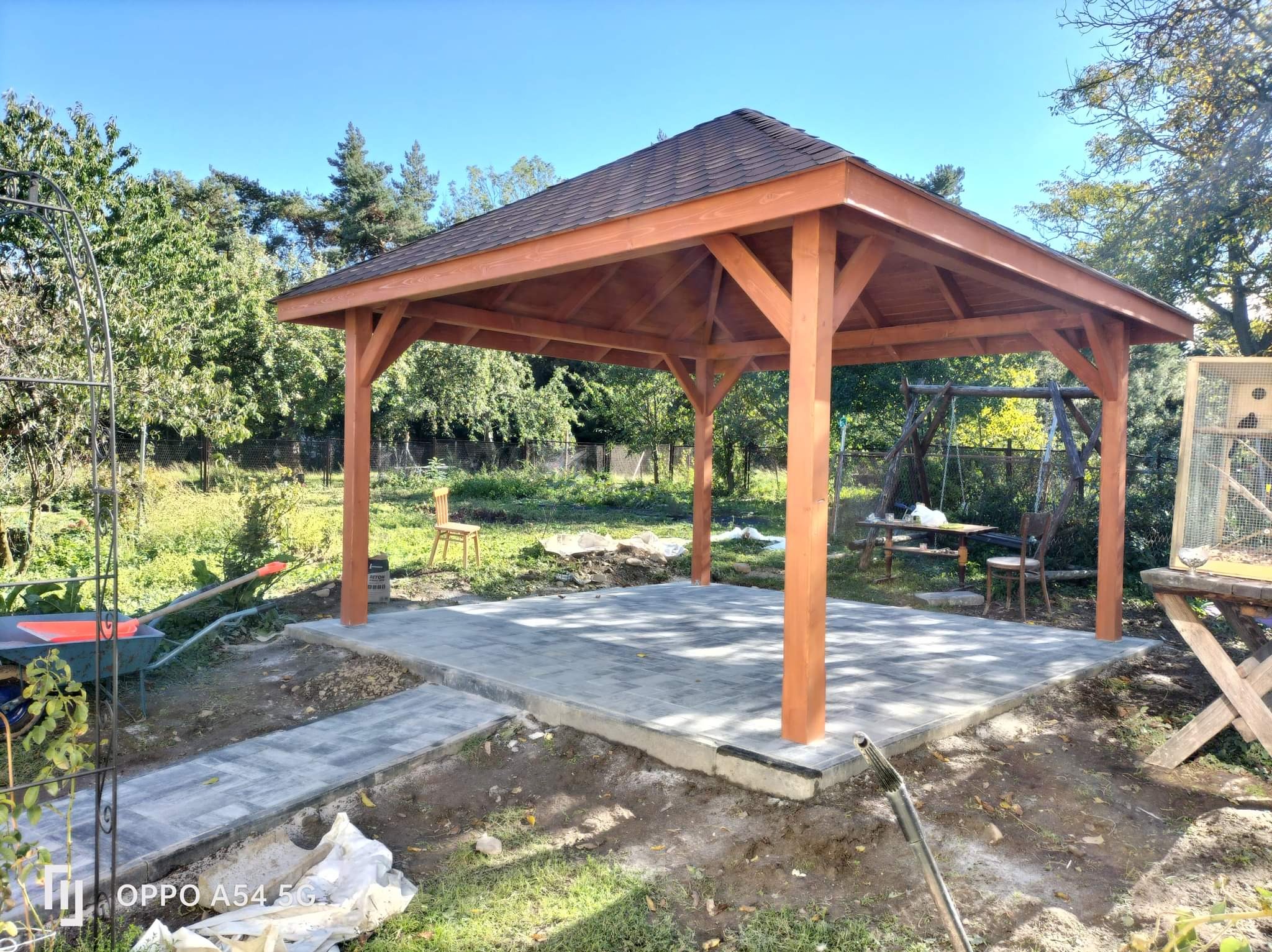 Drewniana altana z brązowym dachem na betonowej podstawie w ogrodzie. Widoczne narzędzia i materiały budowlane. W tle zieleń drzew i krzewów, błękitne niebo.