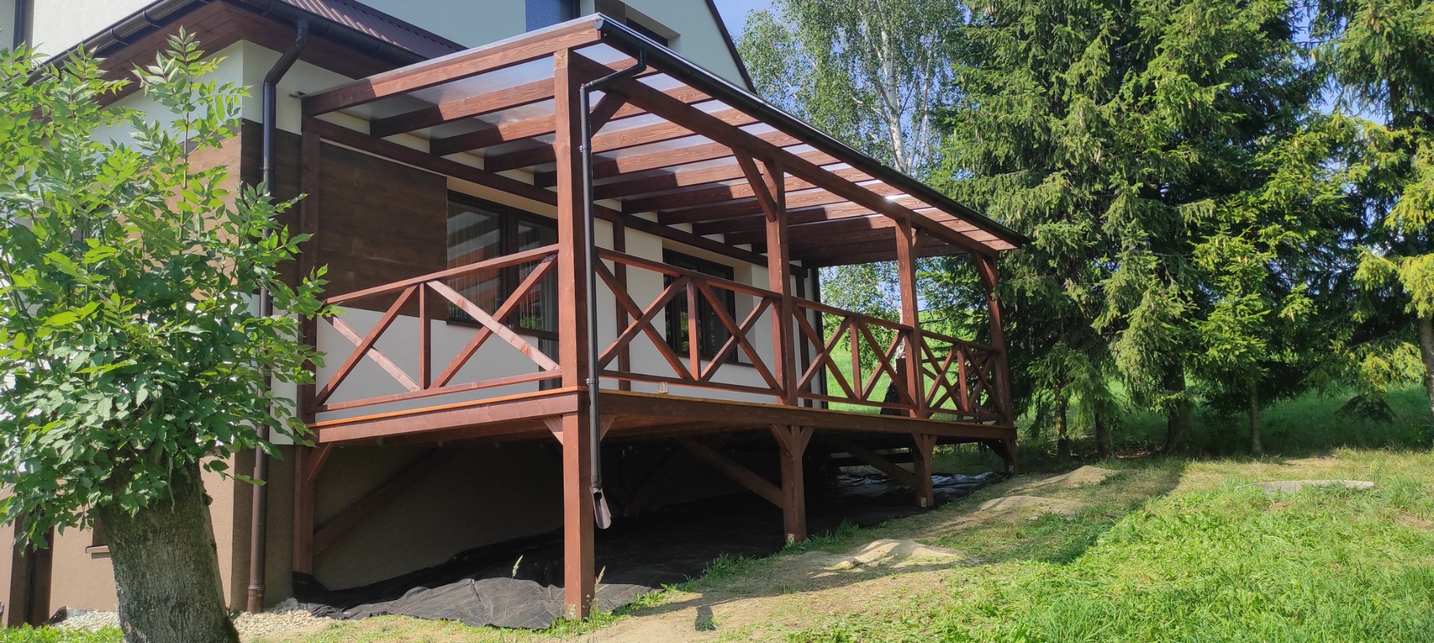 Zadaszony taras z drewnianą balustradą przy domu, widok z boku. Konstrukcja z belek, przezroczyste zadaszenie. W tle zieleń drzew i trawnik.