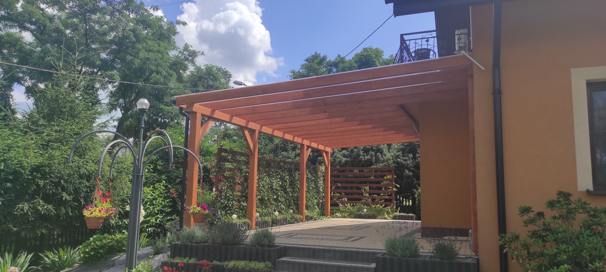 Drewniana pergola przy domu z zadaszeniem z desek, widok z boku, w tle zieleń i błękitne niebo. Oświetlenie ogrodowe i rośliny w donicach na pierwszym planie.