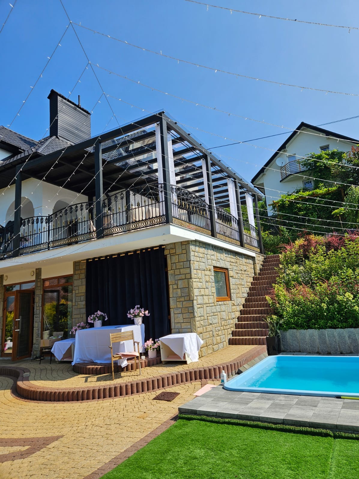 Elegancka pergola z czarną konstrukcją i białymi zasłonami na balkonie. Widok na basen i schody zewnętrzne, dekoracje kwiatowe.