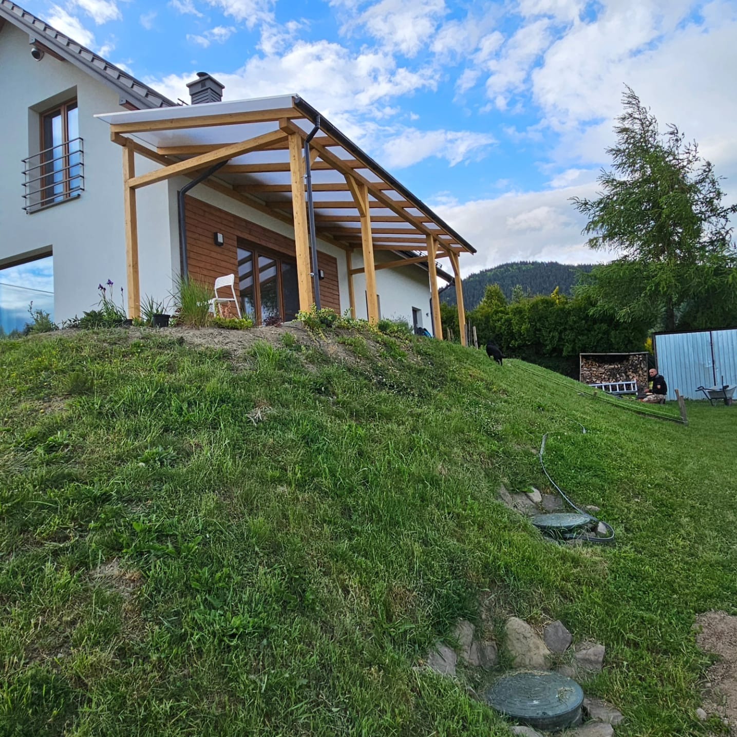 Nowoczesna pergola z drewna i przezroczystego zadaszenia, przylegająca do domu z drewnianymi elementami elewacji. Widok na wzgórze i błękitne niebo.