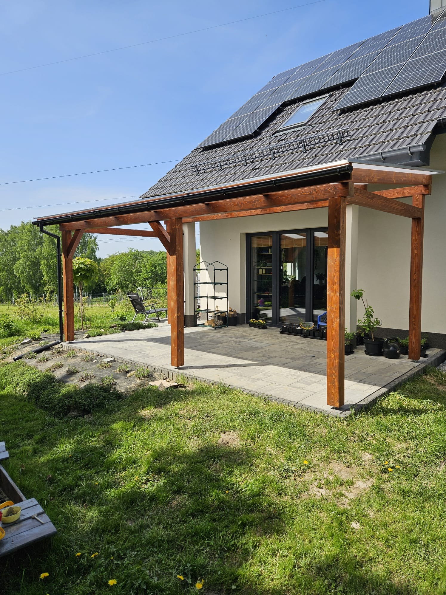 Drewniana konstrukcja zadaszenia tarasu przy domu jednorodzinnym z panelami słonecznymi na dachu. Widok na ogród i elementy wyposażenia.
