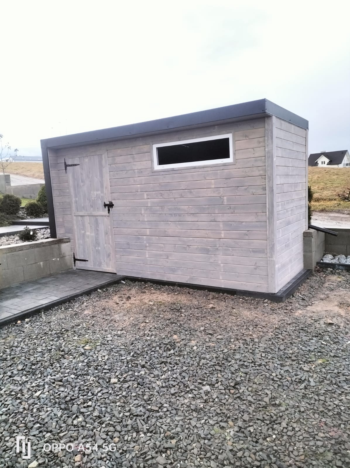 Drewniana altana w odcieniach szarości z oknem i drzwiami, na żwirowym podłożu. Minimalistyczny design, ciemne okucia i obramowania. W tle dom i zieleń.