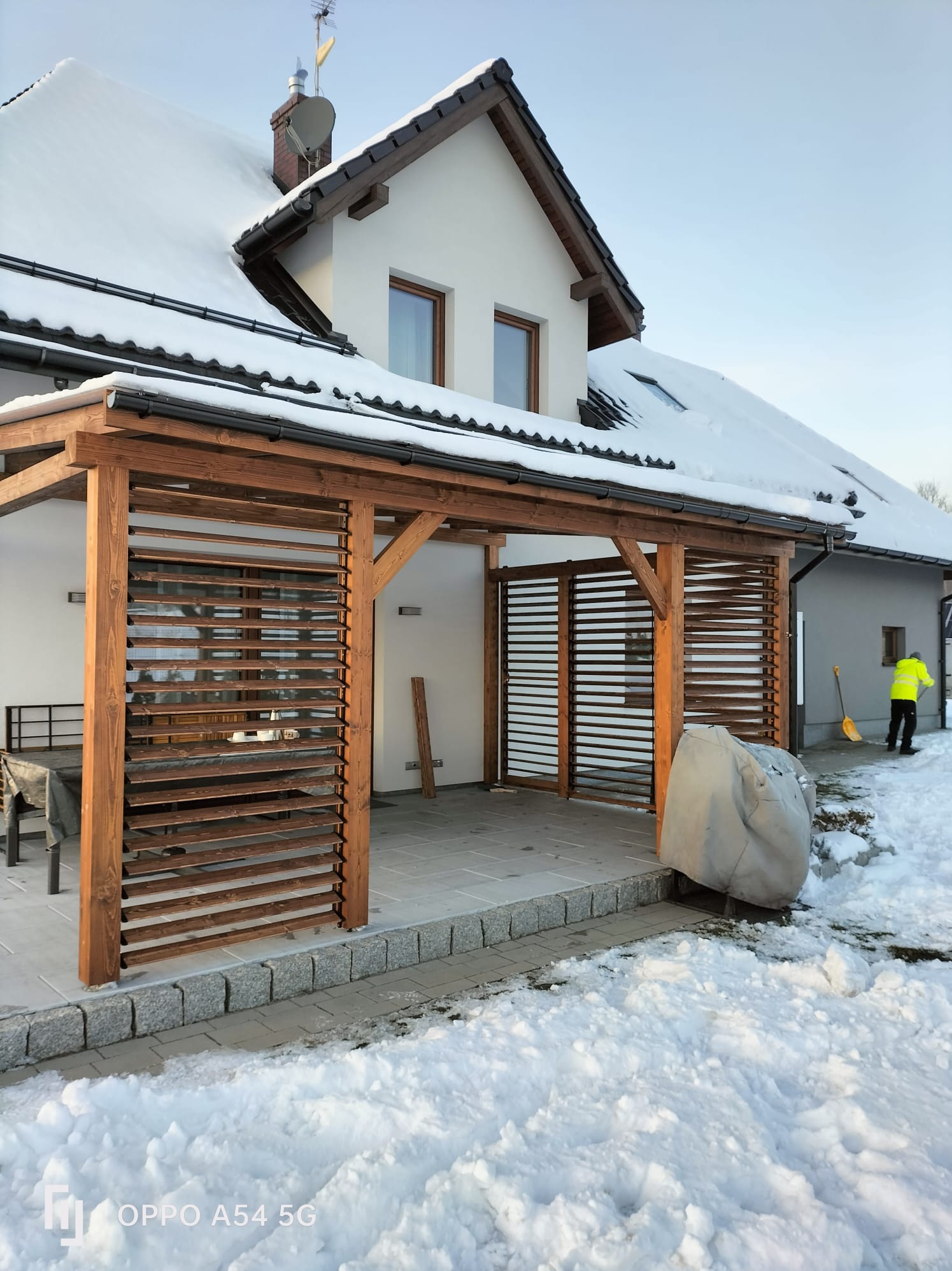 Drewniana pergola z lamelami przy domu, zimowa sceneria. Osoba odśnieża w tle, widoczny zadaszony taras i meble ogrodowe pod przykryciem.