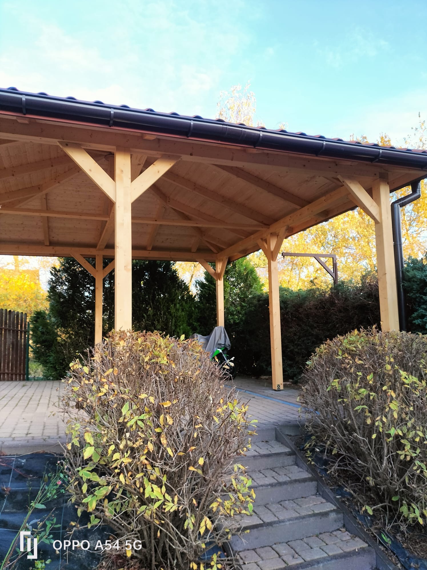 Drewniana konstrukcja z brązowym dachem na zewnątrz, otoczona zielenią i z brukowanym podłożem, widziana z dołu pod kątem.