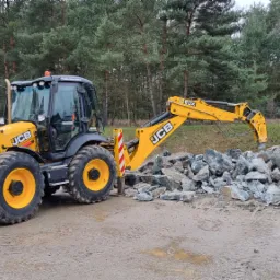Ż&oacute;łto-czarna koparko-ładowarka JCB z zamontowanym młotem hydraulicznym rozbija duży kamień na mniejsze fragmenty na placu budowy, w tle las.