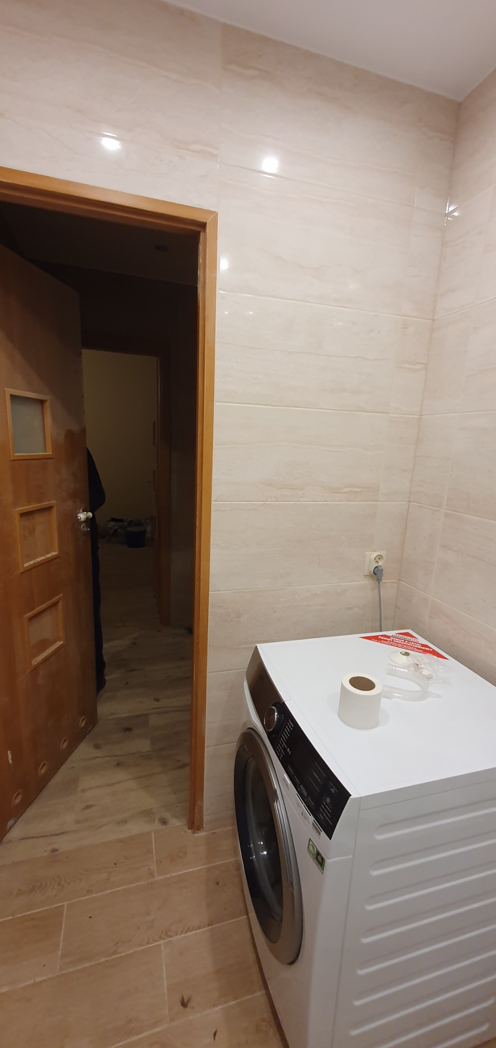 Wykończona łazienka z beżowymi płytkami imitującymi drewno, widoczna pralka z rolką papieru toaletowego i elementami montażowymi na górze, oraz otwarte drewniane drzwi do kolejnego pomieszczenia.