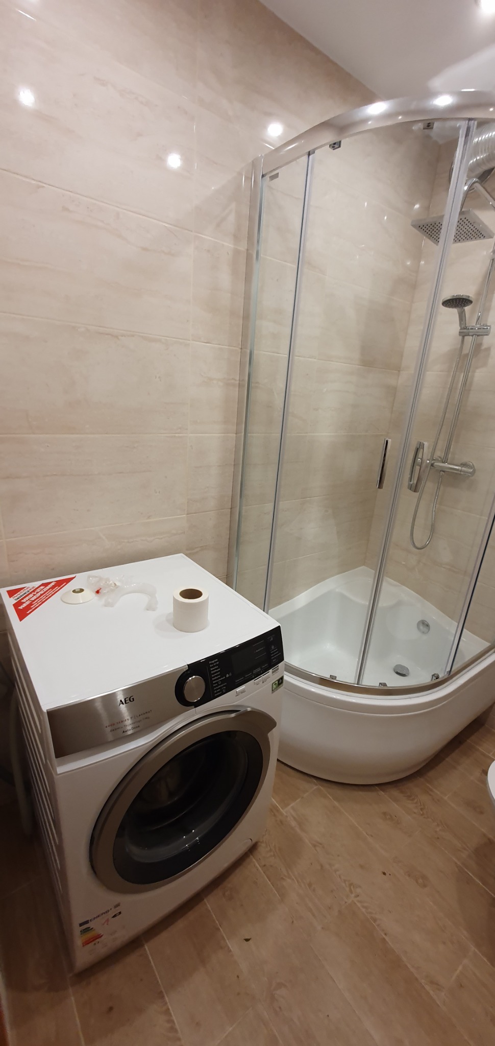 Nowa pralka AEG obok kabiny prysznicowej w łazience wykończonej beżowymi płytkami, na pralce leżą resztki opakowania i rolka papieru toaletowego.