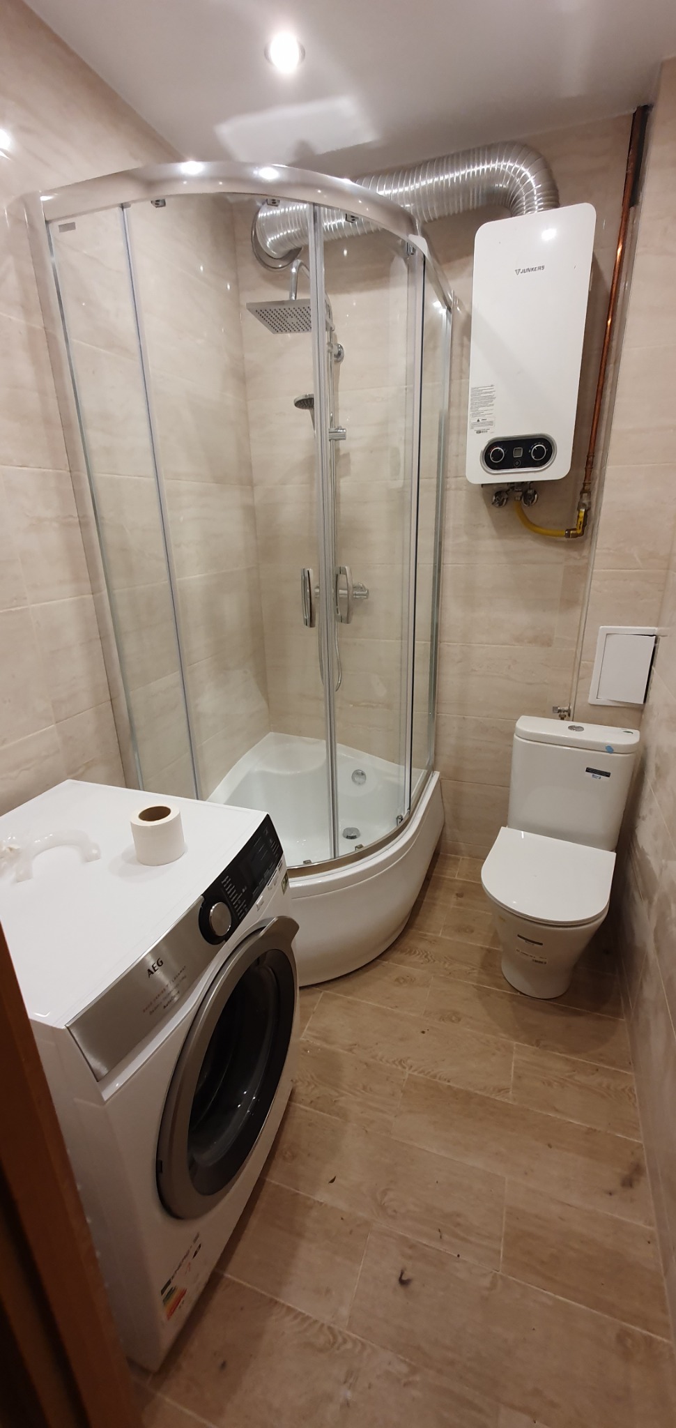 Wykończona łazienka z kabiną prysznicową narożną, pralką AEG i piecykiem Junkers, wykończona płytkami imitującymi drewno
