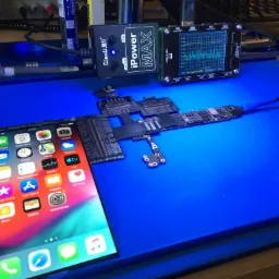 Rozłożony iPhone X na niebieskiej macie serwisowej obok miernika napięcia i prądu iPower MAX, z widocznym ekranem głównym i narzędziami serwisowymi w tle.