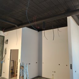 Volodymyr Spichak - Wnętrze w trakcie remontu: białe ściany po szpachlowaniu i malowaniu, czarny sufit z widoczną instalacją, drabina malarska i narzędzia na podłodze.