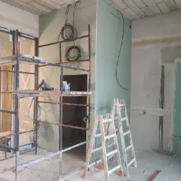 Wnętrze w trakcie prac: metalowe rusztowanie z narzędziami, dwie drewniane drabiny oparte o ścianę z płyt, widoczne kable elektryczne i sufit z wykończeniem przypominającym cegłę.
