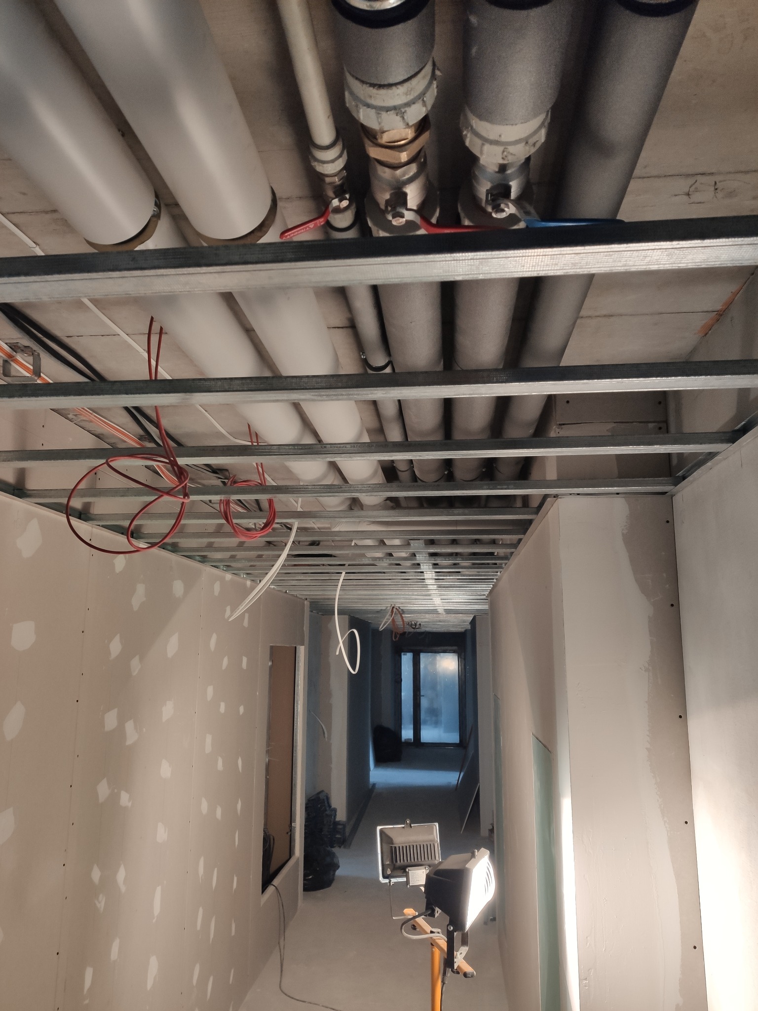 Korytarz w trakcie remontu, widoczne profile sufitowe, instalacje pod sufitem, ściany przygotowane do dalszych prac, oświetlenie robocze na statywie.