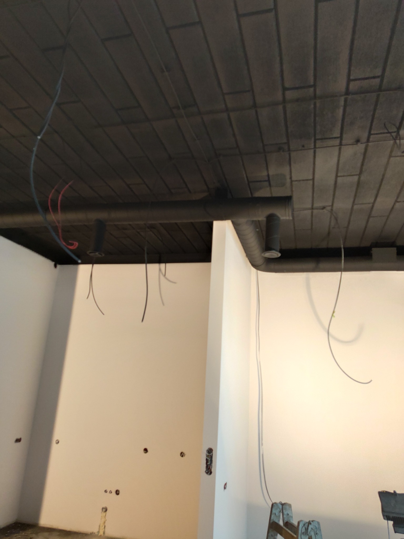 Szary sufit z widoczną instalacją wentylacyjną i wiszącymi kablami elektrycznymi w pomieszczeniu z białymi ścianami w trakcie prac wykończeniowych.