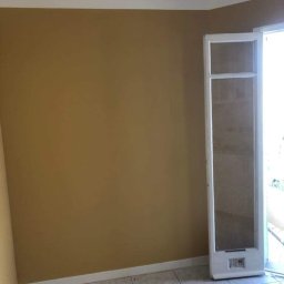 NewTomorrowHome - Pokój po malowaniu ścian na musztardowy kolor, widoczna biała listwa przypodłogowa, fragment okna i płytek podłogowych.