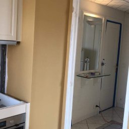 NewTomorrowHome - Ściana w kuchni pomalowana na żółto-złoty kolor, widoczna dziura w ścianie obok szafki, fragment piekarnika i przejście do korytarza z płytkami na podłodze.