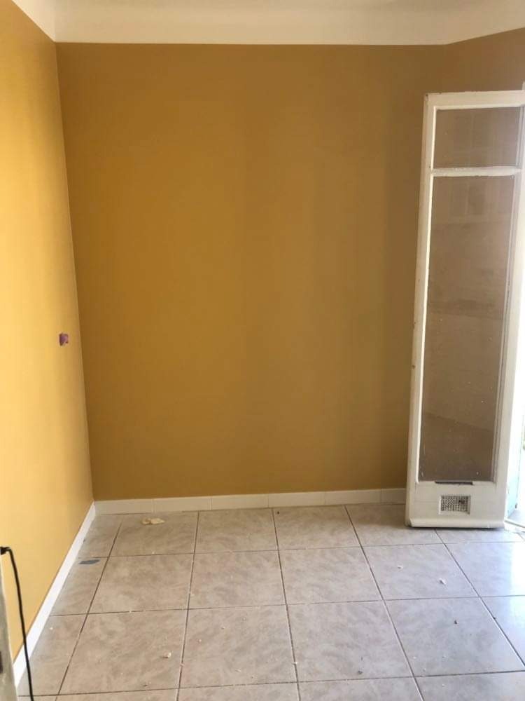 Pomalowane na musztardowy kolor ściany w pustym pokoju z beżową podłogą z płytek; widoczny biały grzejnik ścienny i biała listwa przypodłogowa.