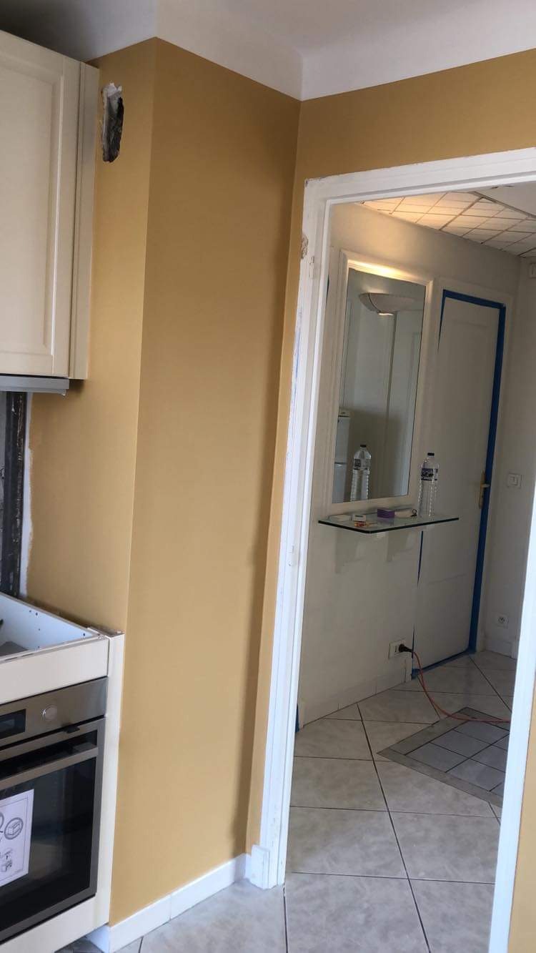 Ściana w kuchni pomalowana na żółto-złoty kolor, widoczna dziura w ścianie obok szafki, fragment piekarnika i przejście do korytarza z płytkami na podłodze.