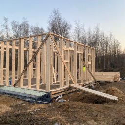Łukasz Bęc - Konstrukcja szkieletowa domu w trakcie budowy, widoczne drewniane belki i słupy na fundamencie, w tle drzewa bez liści i hałda desek.