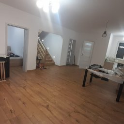 Łukasz Bęc - Przestronne wnętrze domu w trakcie prac wykończeniowych: drewniana podłoga, białe ściany, schody z oświetleniem punktowym, widoczny stół roboczy i zabezpieczenie dla dziecka przy komodzie.