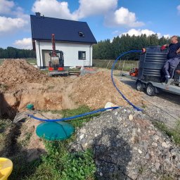 Łukasz Bęc - Montaż szamba betonowego na posesji z użyciem mini koparki i specjalistycznego sprzętu do transportu i montażu, widoczny pracownik w odzieży roboczej.