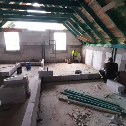 Łukasz Bęc - Wnętrze poddasza w trakcie murowania ścian z bloczków, widoczny pracownik w stroju roboczym, narzędzia i materiały budowlane, surowa konstrukcja dachu z drewnianych belek.