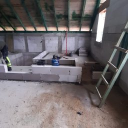 Łukasz Bęc - Wnętrze poddasza z częściowo wymurowanymi ścianami z białych bloczków, widoczne narzędzia murarskie, drabina oparta o ścianę, surowy drewniany strop.