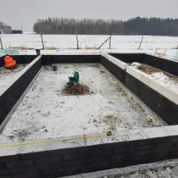 Fundament domu murowanego w trakcie budowy, widoczne bloczki betonowe i warstwa izolacji, dwóch pracowników w odblaskowych ubraniach, zimowy krajobraz w tle.