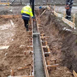 Wylewanie betonu do szalunku zbrojeniowego pod fundament w zimowej scenerii, pracownik w odblaskowej kamizelce nadzoruje proces, w tle ośnieżone drzewa.