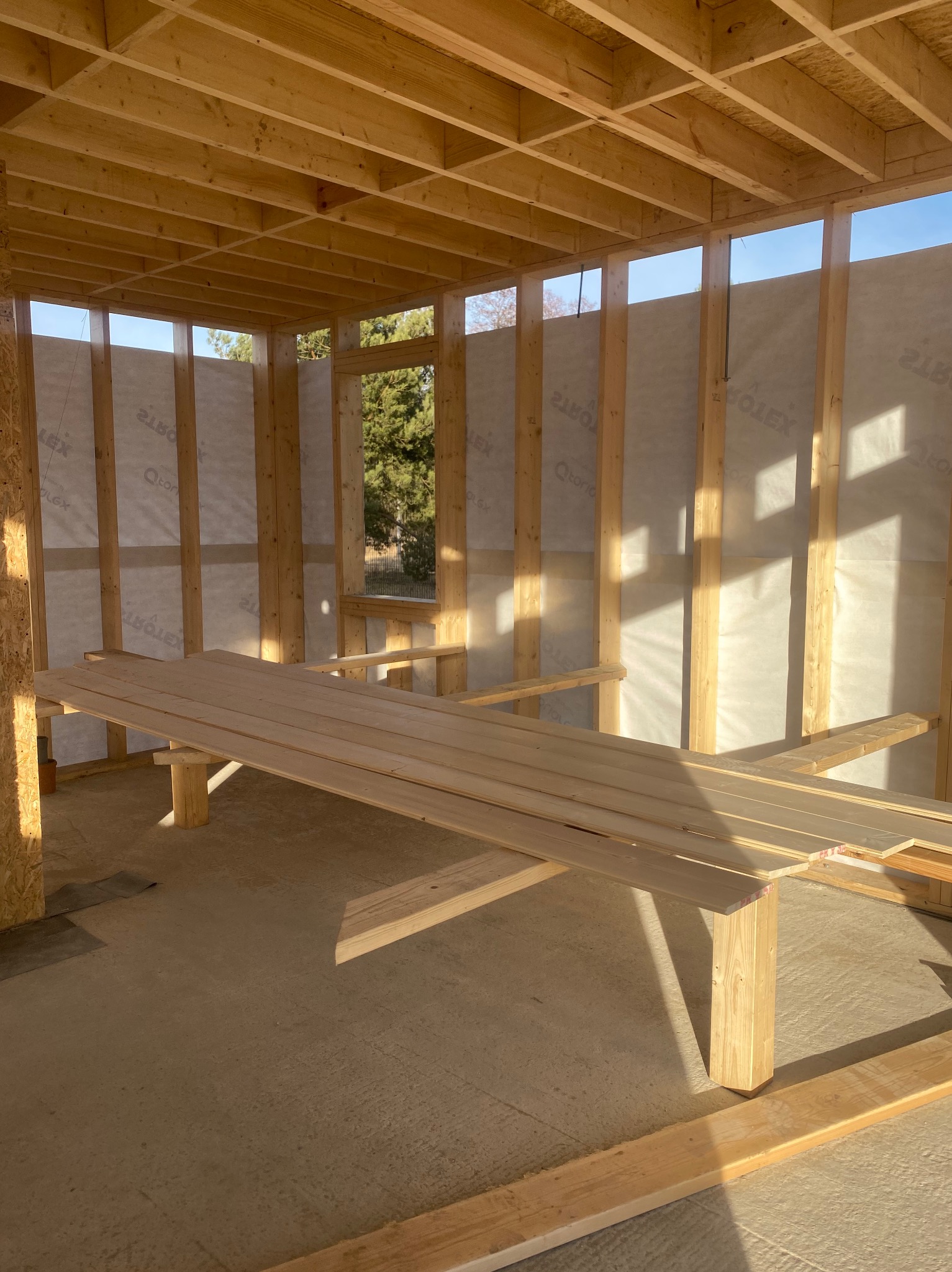 Surowy szkielet drewniany domu w trakcie budowy, z widocznym oknem i deskami przygotowanymi do montażu, oświetlony porannym słońcem.