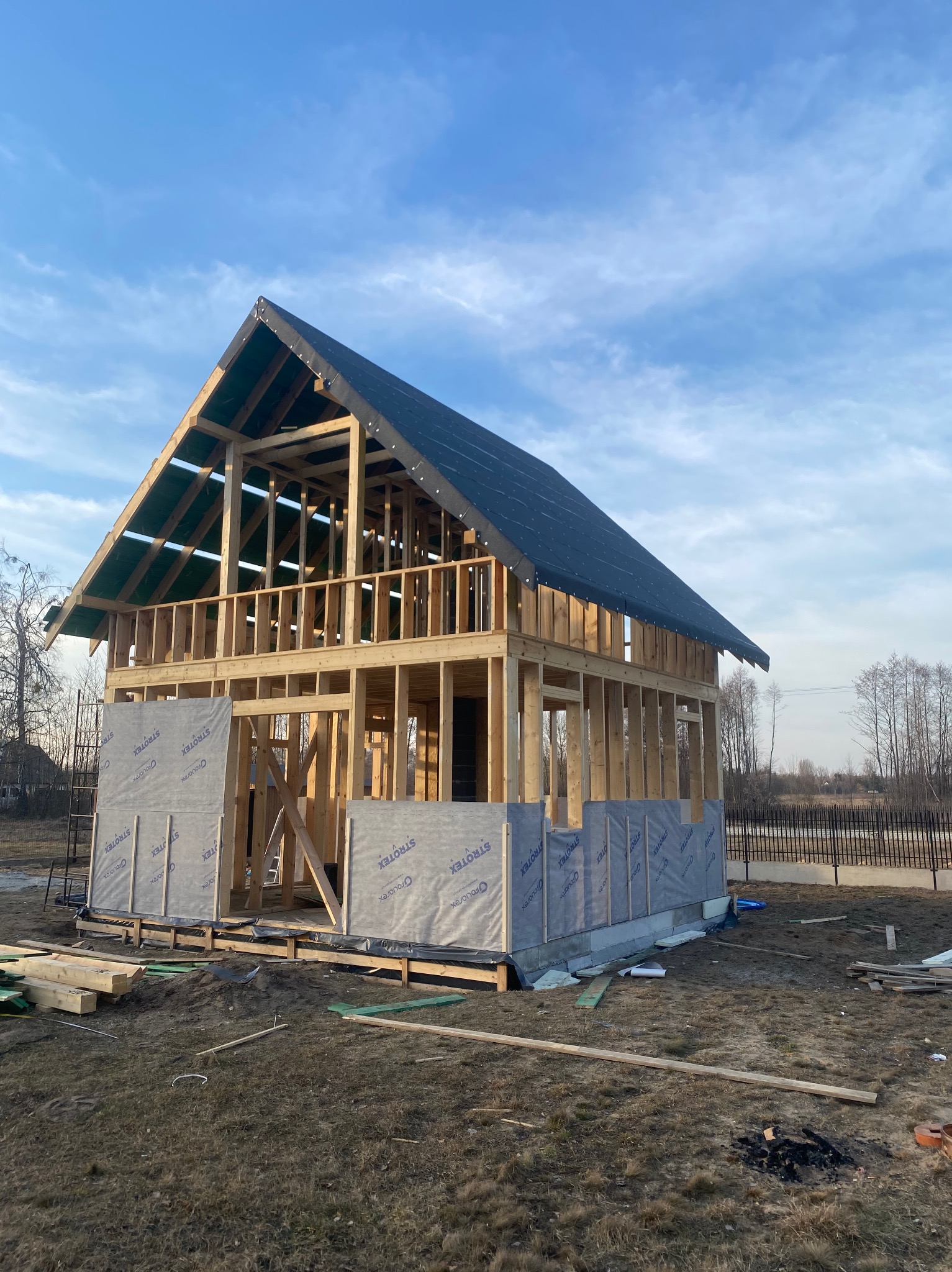 Surowy szkielet drewnianego domu w trakcie budowy, widoczne membrany izolacyjne na fundamentach, błękitne niebo w tle.