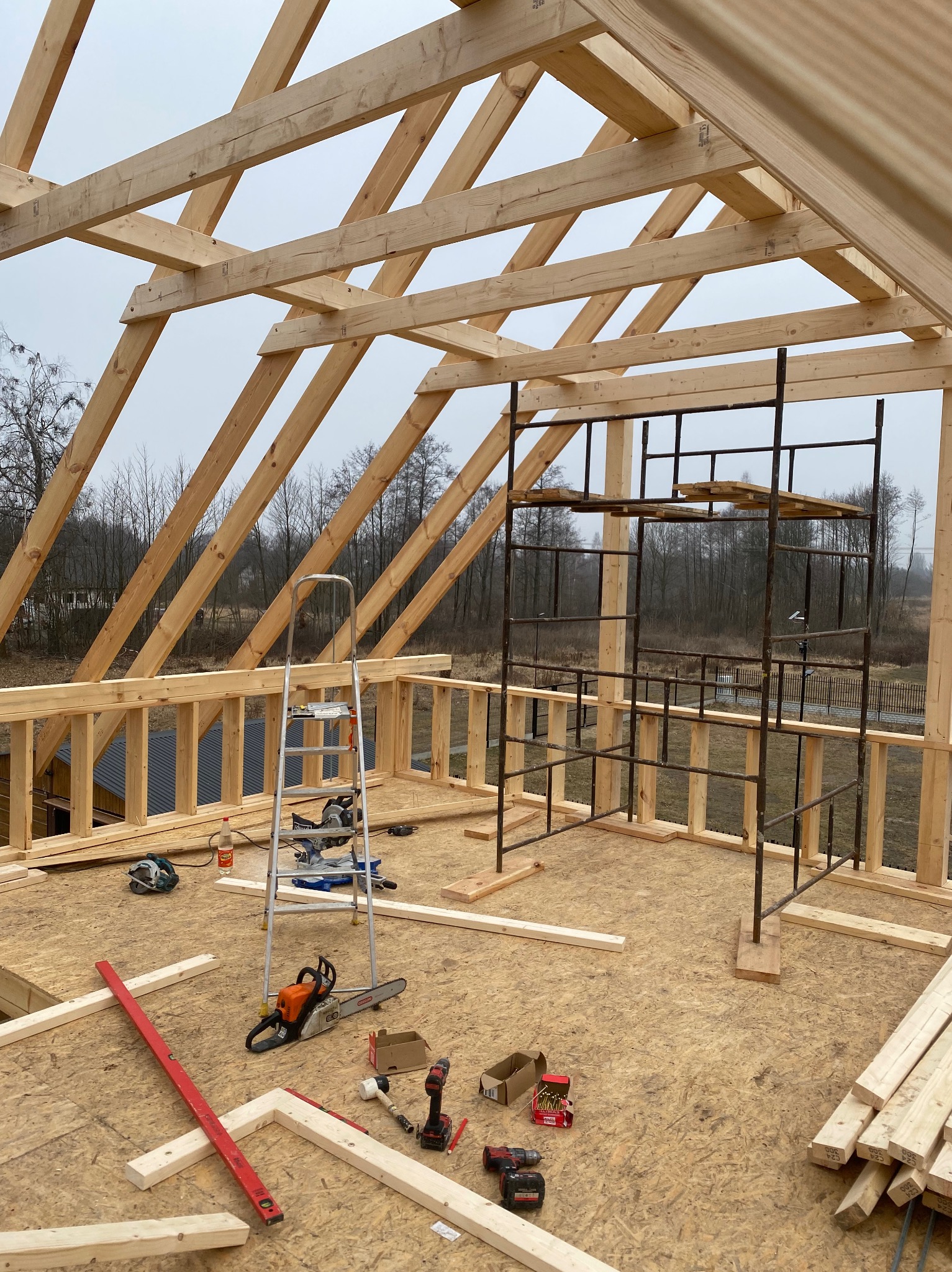 Konstrukcja drewniana dachu w trakcie budowy, widoczne krokwie, narzędzia takie jak piła łańcuchowa, poziomica i wiertarka na podłodze z płyty OSB, w tle drzewa i szare niebo.