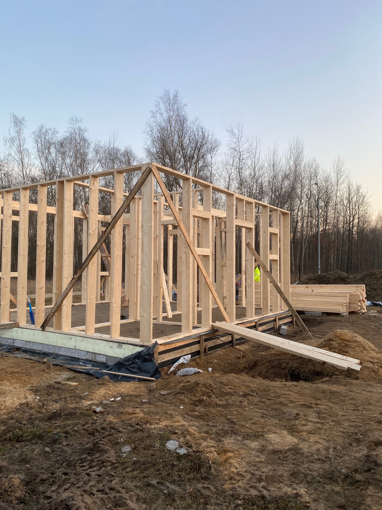 Konstrukcja szkieletowa domu w trakcie budowy, widoczne drewniane belki i słupy na fundamencie, w tle drzewa bez liści i hałda desek.