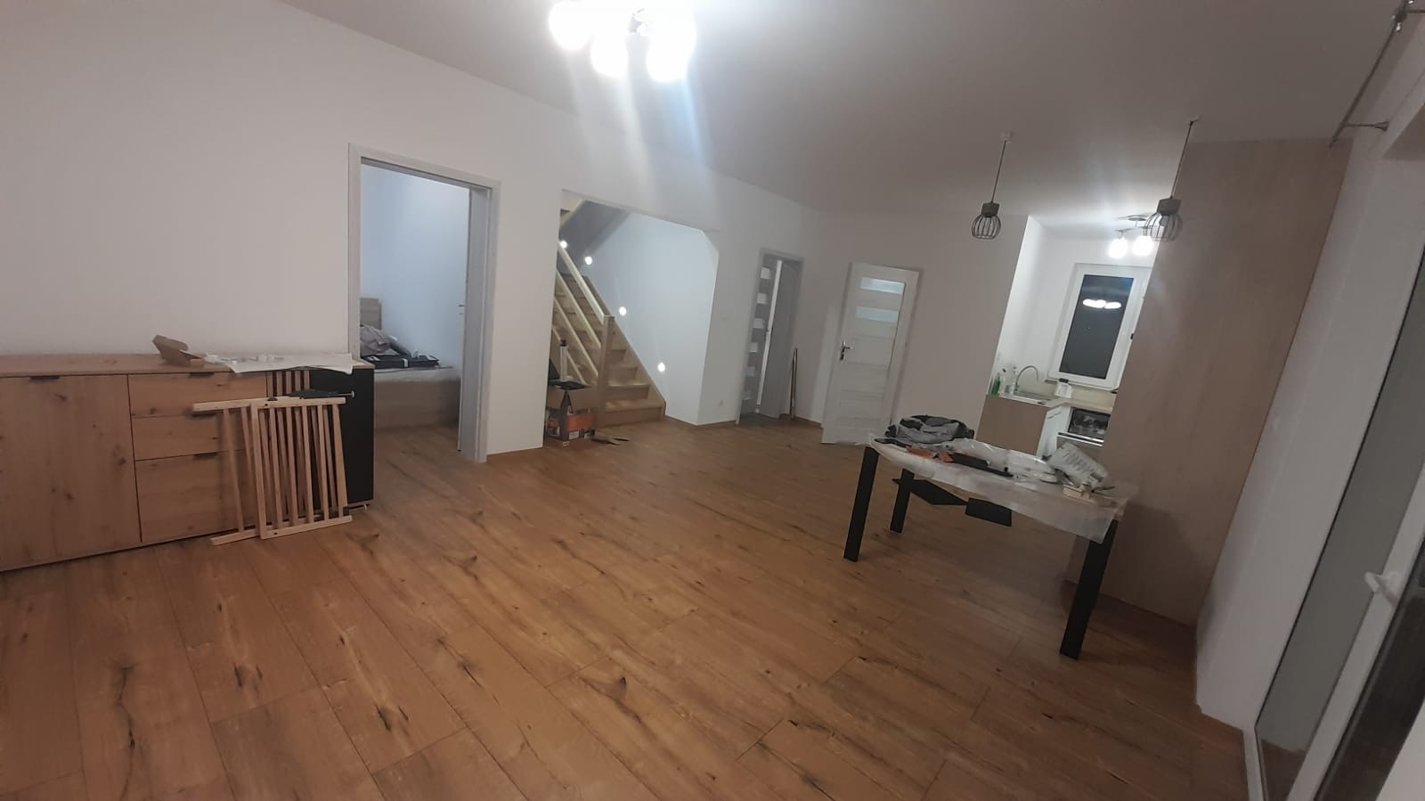Przestronne wnętrze domu w trakcie prac wykończeniowych: drewniana podłoga, białe ściany, schody z oświetleniem punktowym, widoczny stół roboczy i zabezpieczenie dla dziecka przy komodzie.