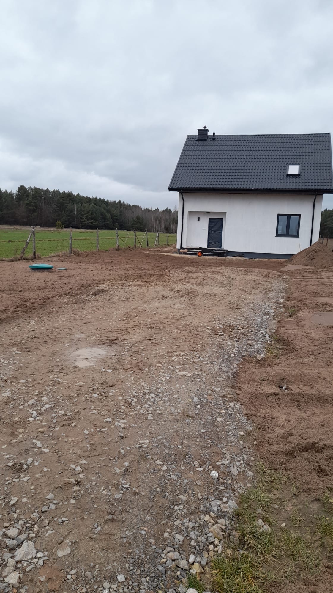 Nowo wybudowany, biały dom z ciemną dachówką w surowym otoczeniu placu budowy, widoczne ślady prac ziemnych i materiały budowlane.