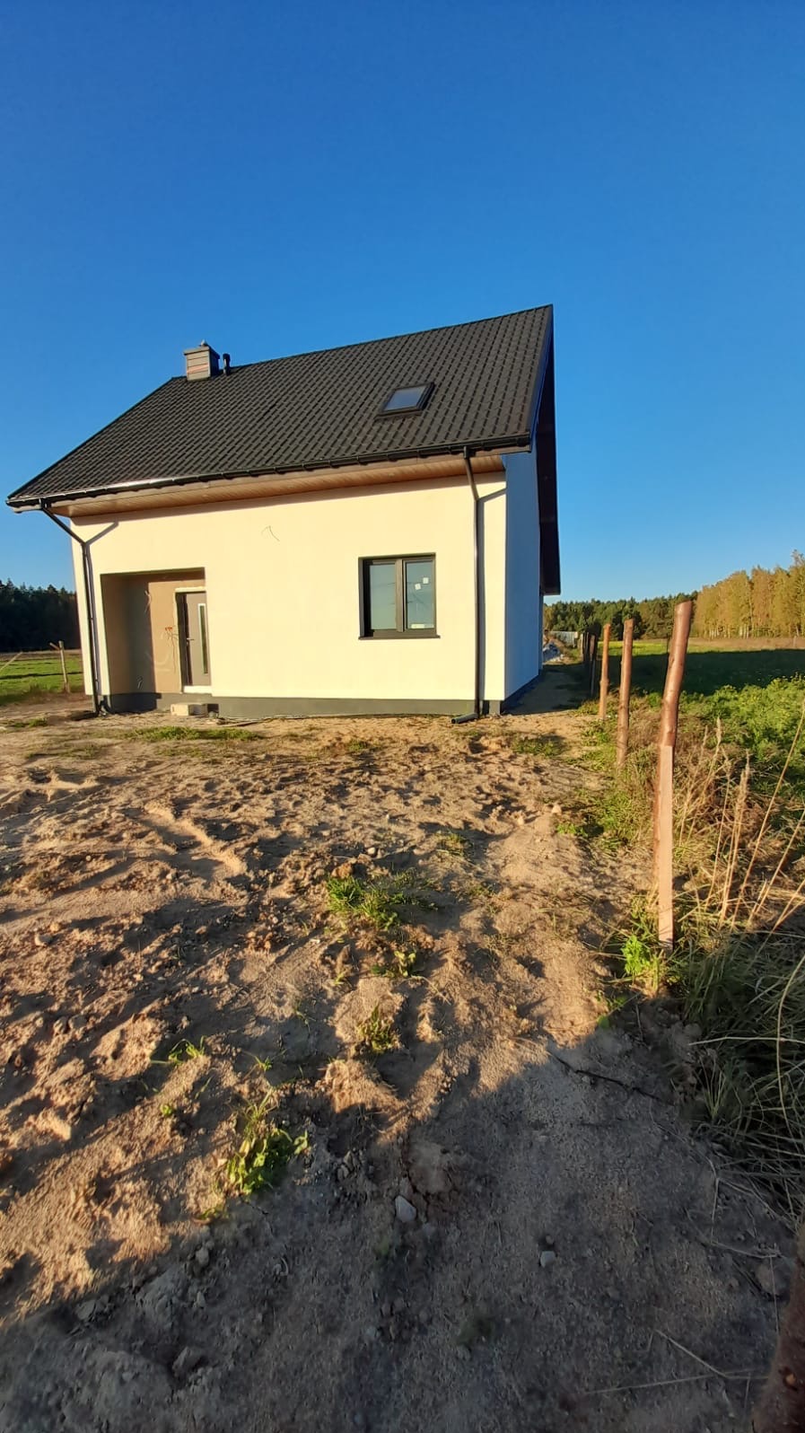 Nowoczesny, parterowy dom z jasną elewacją, ciemnym dachem i pojedynczym oknem, widoczny z zewnątrz, z surowym otoczeniem i częściowo postawionym płotem.