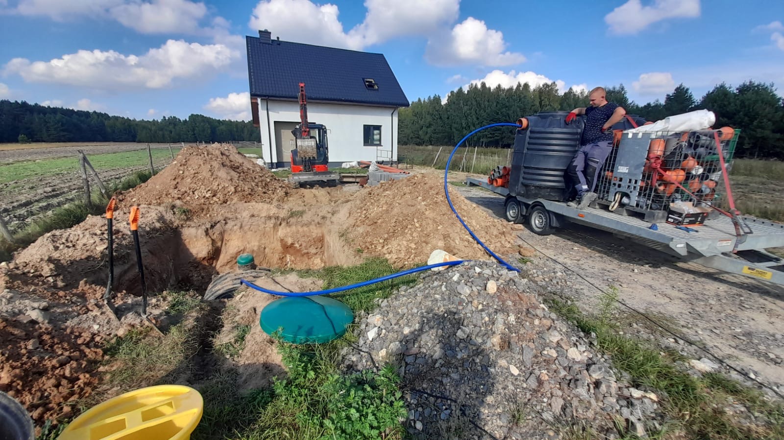 Montaż szamba betonowego na posesji z użyciem mini koparki i specjalistycznego sprzętu do transportu i montażu, widoczny pracownik w odzieży roboczej.