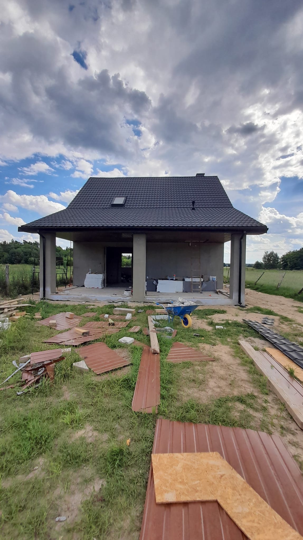 Surowy stan domu murowanego z czarną dachówką, widoczne materiały budowlane i taczka przed budynkiem, tło stanowi zachmurzone niebo i zieleń.