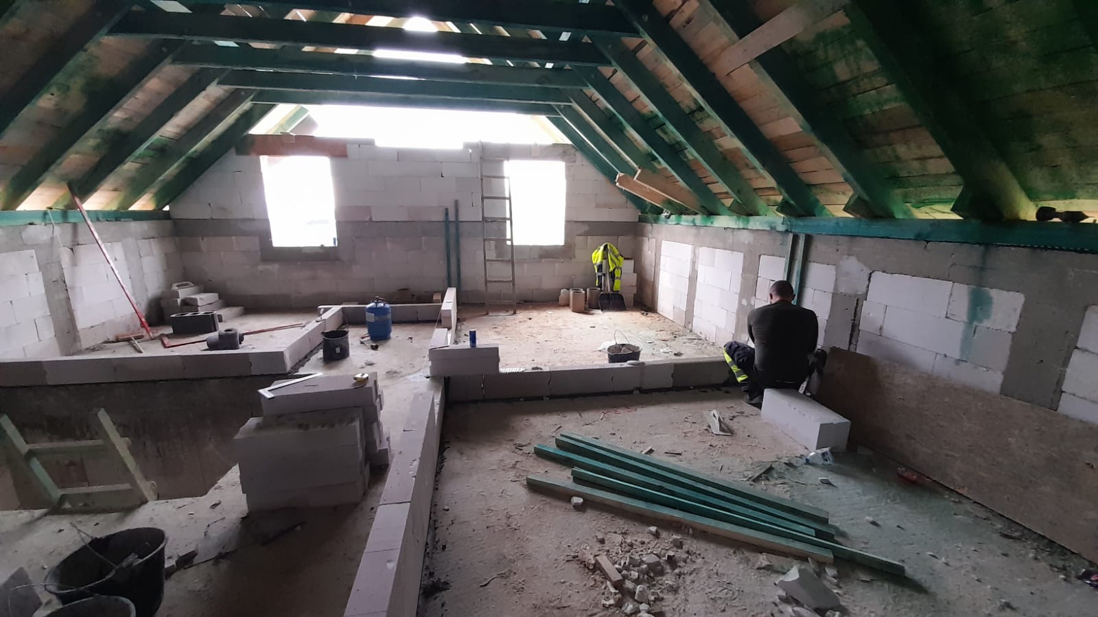 Wnętrze poddasza w trakcie murowania ścian z bloczków, widoczny pracownik w stroju roboczym, narzędzia i materiały budowlane, surowa konstrukcja dachu z drewnianych belek.