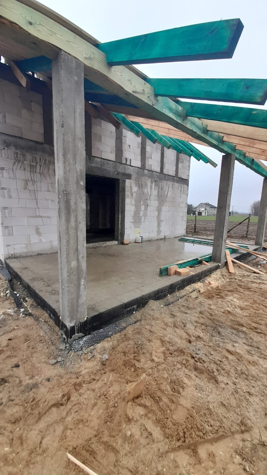 Surowy stan budowy domu z betonu komórkowego: wylana podłoga, betonowe słupy, drewniana konstrukcja dachu pomalowana na zielono, widoczna izolacja fundamentów.