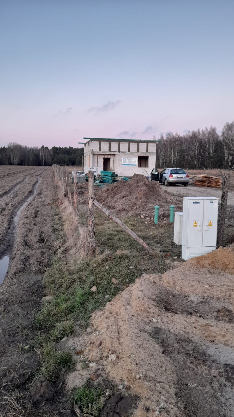 Surowy stan domu w trakcie budowy z widocznymi pustakami, stertą ziemi, skrzynką elektryczną i zaparkowanym samochodem na tle pola i lasu.