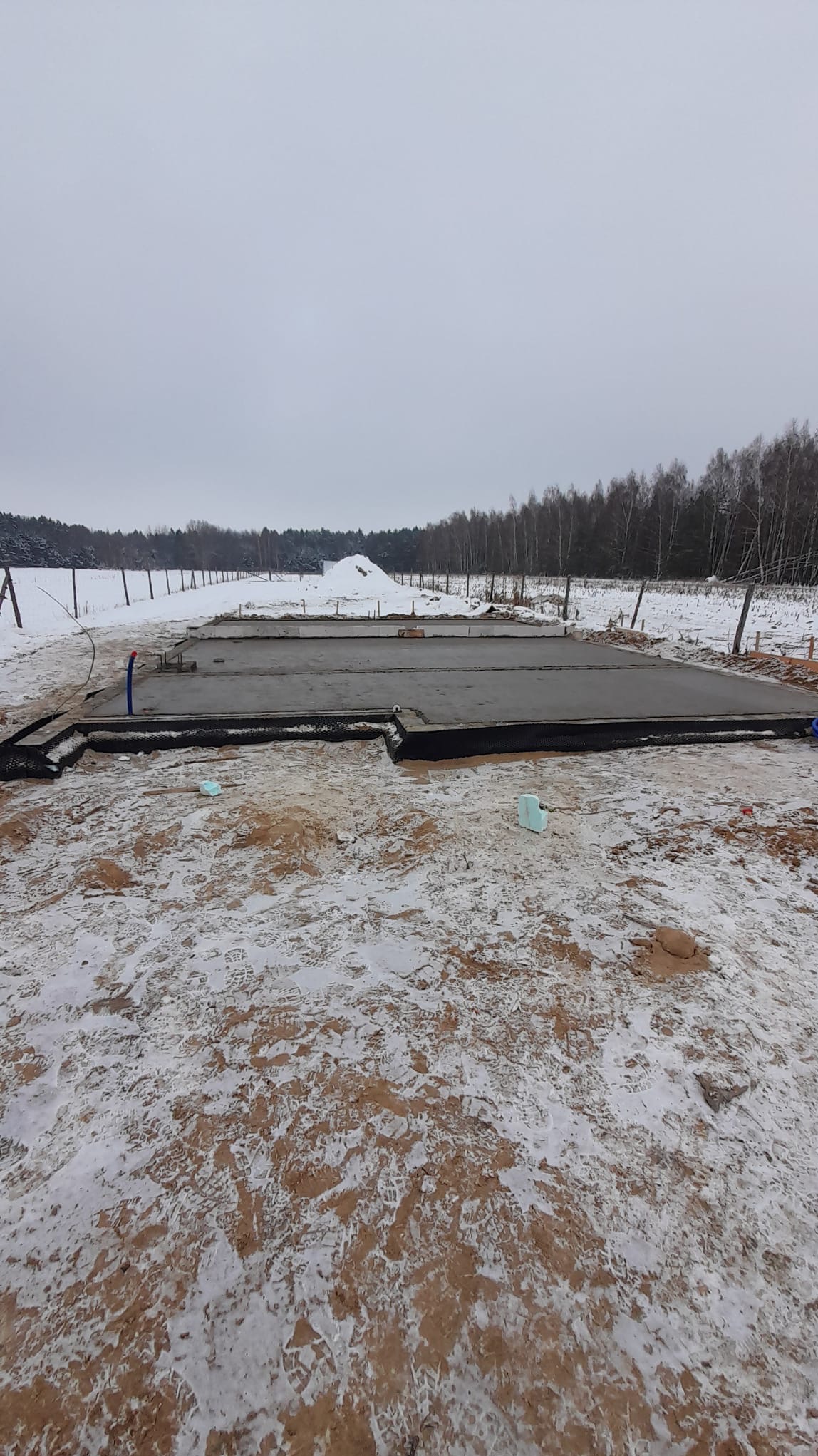 Świeżo wylany fundament pod dom jednorodzinny, widok na zasypany śniegiem teren budowy i las w tle.