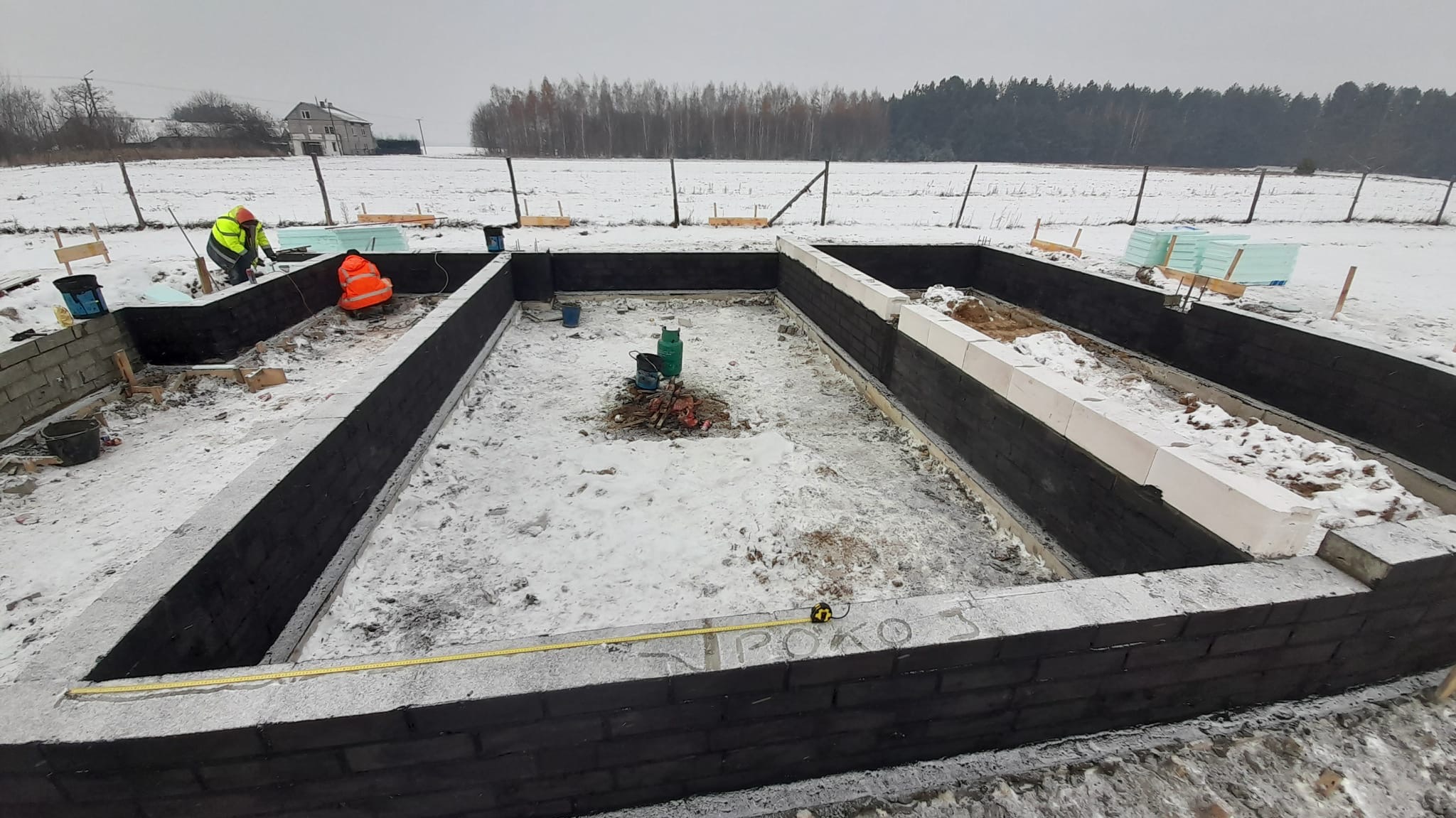 Fundament domu murowanego w trakcie budowy, widoczne bloczki betonowe i warstwa izolacji, dwóch pracowników w odblaskowych ubraniach, zimowy krajobraz w tle.