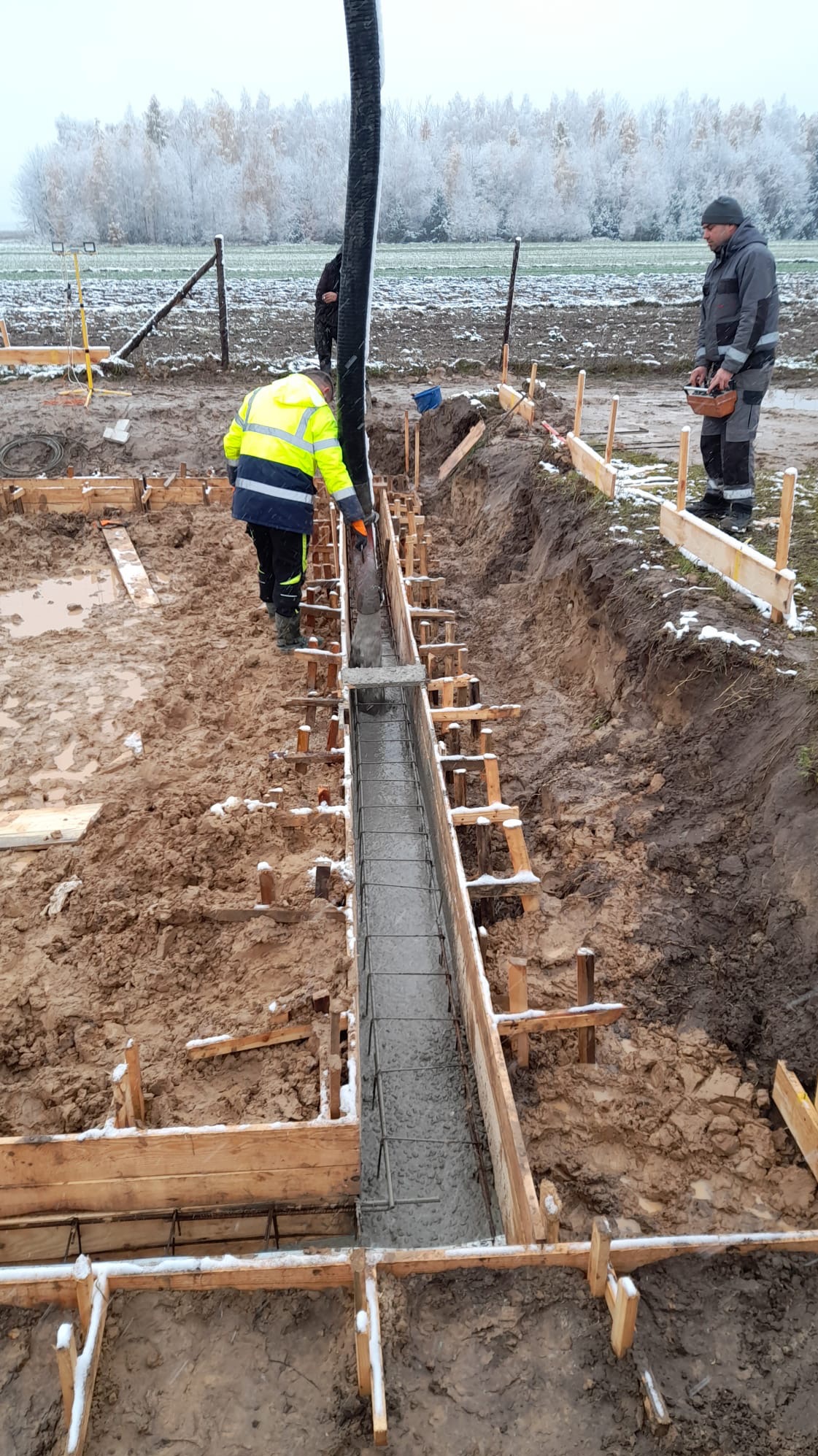 Wylewanie betonu do szalunku zbrojeniowego pod fundament w zimowej scenerii, pracownik w odblaskowej kamizelce nadzoruje proces, w tle ośnieżone drzewa.