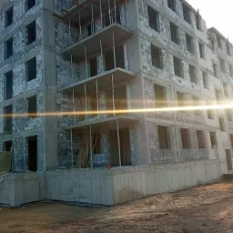 Wielopiętrowy budynek w trakcie budowy z szarych bloczków, widoczne rusztowania i podpory stropowe, teren budowy z ziemią.